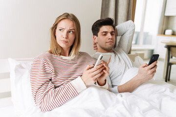 Obraz na płótnie Canvas Portrait of displeased couple using mobile phones while lying in bed