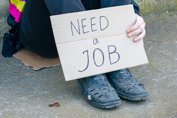 unemployed in old shoes holds in his hand a cardboard tablet with the inscription need a job on the...