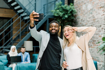 Happy business people, working in office. Smiling Caucasian lady and African man making joint selfie photo on smartphone, posing, laughing and showing dofferent gestures