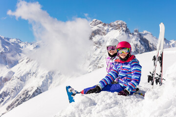 Fototapeta na wymiar Two happy girls sit in the mountain with ski over high peaks and clouds in snow look at camera smiling
