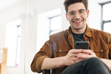 Image of joyful young man winking and using cellphone while working