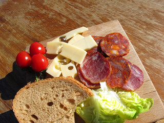 Homemade sausage, salami, cheese, cherry tomatoes, lettuce and bread on a wooden board. Simple and delicious food on an old wooden table lit by the bright sun