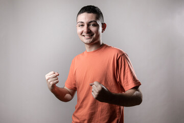 Ragazzo giovane con t-shirt arancione fa un gesto di esultanza , isolato su sfondo grigio