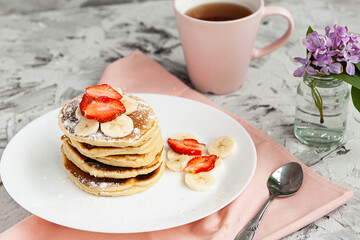 A stack of pancakes with a pink tea mug and  fresh strawberries on a white plate on a light background. A small bouquet of lilacs in a glass vase stands next to the Breakfast.