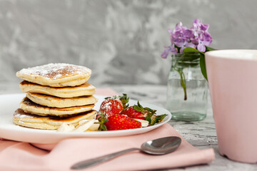 A stack of pancakes with a pink tea mug and  fresh strawberries on a white plate on a light background. A small bouquet of lilacs in a glass vase stands next to the Breakfast.