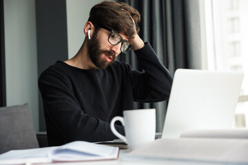 Image of young confused man using wireless earphones and working with laptop