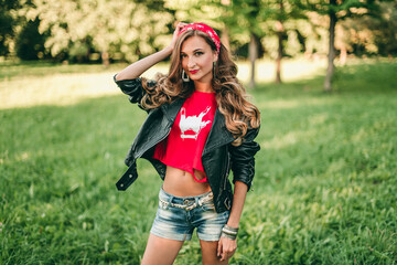 Portrait of beautiful young blonde stylish woman with long curly hair and red lipstick in rock style in short denim shorts, red headband, t-shirt, black leather jacket.