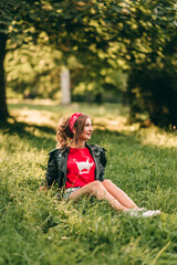 Fototapeta premium Portrait of beautiful young blonde stylish woman with long curly hair and red lipstick sitting on the green grass in rock style in short denim shorts, red headband, t-shirt, black leather jacket.