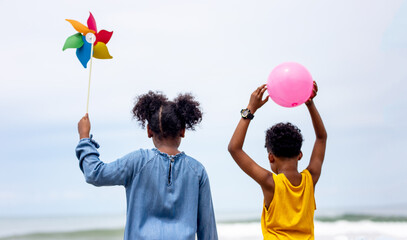Rear view of Young kids holding color balloons and rainbow windmill walking on beach.