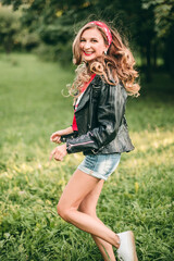 Fototapeta premium Portrait of beautiful young blonde stylish woman with long curly hair and red lipstick sitting on the green grass in rock style in short denim shorts, red headband, t-shirt, black leather jacket.
