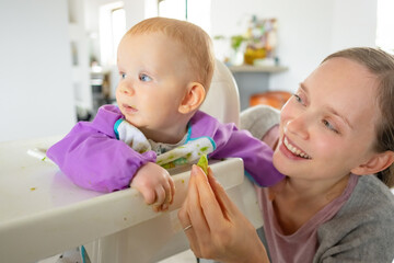 Joyful mom feeding baby daughter in high chair, giving piece of fresh green vegetable. Closeup shot. Childhood or child care at home concept