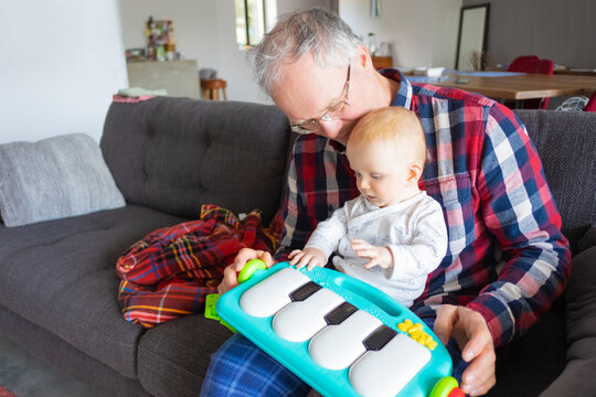 Grey haired grandfather sitting on couch and playing with baby granddaughter and her toy piano. Family or creative skills concept