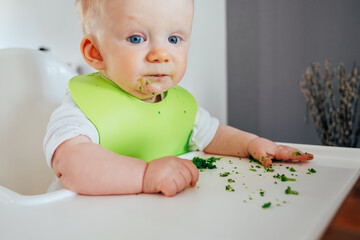 Portrait of cute baby girl sitting messy after feeding. Little child chewing at dining table. Led weaning and self-feeding concept