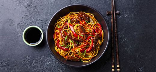 Stir fry noodles with vegetables and beef in black bowl. Slate background. Top view.