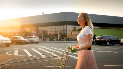 Beautiful smiling blonde girl pushing shopping cart while walking on car parking at shopping mall © Kyrylo Ryzhov