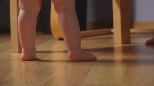 Low-angle feet view of little adorable baby standing on the tiptoes next to the chair