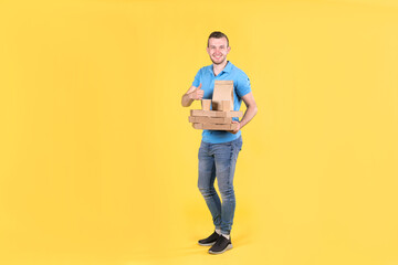 Young smiling guy delivering food from restaurant in blue uniform with thermo bag on his back holds an order for customer on yellow studio background. Fast food delivery to office or home