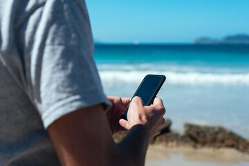 Young man typing a message on his phone on the beach