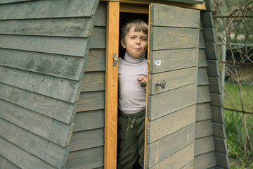 Beautiful little boy with blond hair looks out smiling from the door of a gray house