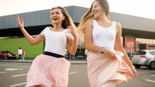 Portrait Of Two Laughing Cheerful Girls In Same Outfit Playing And Having Fun On Car Parking At Shopping Mall