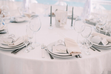 Restaurant, banquet, table setting. The table is covered with a white tablecloth, dishes, white clean plates, cutlery, napkins, wine glasses, candles. Close-up.