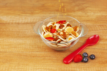 Mini pancakes with strawberries and blueberries in a bowl on a wooden table, top view. Trendy food - pancake cereal