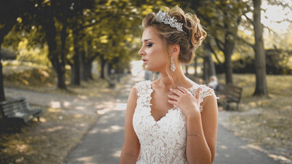 Toned portrait of elegant blonde bride in white dress posing on long alley at park