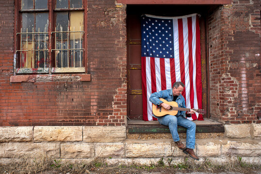 Man wearing cowboy hat, playing acoustic guitar outside sitting on old factory loading dock in city with American flag hanging