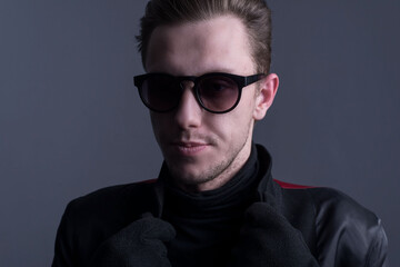 Close-up studio portrait of a twenty-five-year-old young man in dark clothes and sunglasses, holding himself by the collar in black gloves. On a gray background.