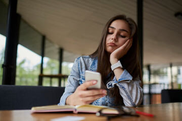 Exhausted young woman browsing internet stores on smartphone in coffee shop