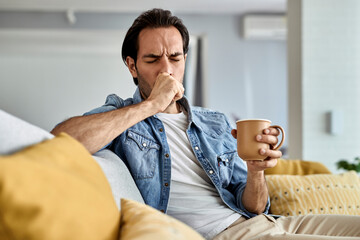 Young man coughing while drinking tea in the living room.