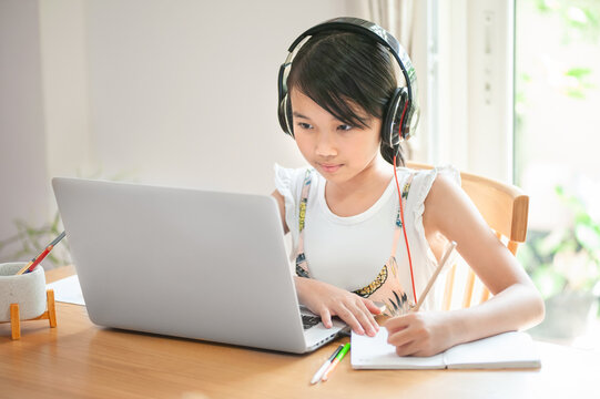 little asian girl sitting at home at a table using laptop and  headphone to working  and writing on her homework Homeschool for social distancing concept
