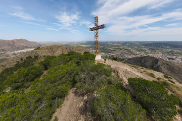 An aerial view with the drone from the summit cross of the mountain Muela north of the eastern Spanish city of Orihuela. A male hiker stands in front of the cross. It is a sunny day in June with cloud