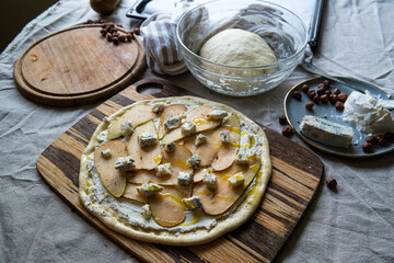 Step by step process of making pizza with pear, ricotta and gorgonzola. Adding gorgonzola cheese and olive oil on the top of pizza on wooden board. Bowl with dough, hazelnuts and cheese on background.