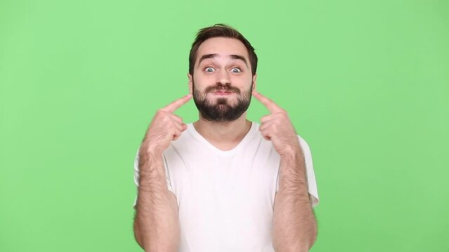 Fun young guy 20s in white t-shirt isolated on green chroma key background studio. People emotions lifestyle concept. Looking camera fooling around pointing index fingers on blowing cheeks monkey ears