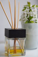 close-up glass diffuser with a black cap, wooden sticks and yellow aromatic oil. white wall and white glass with white flowers in the background. selective focus