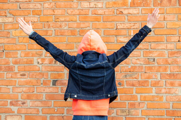 Young girl stands with her back and holds her hands up on the background of a brick red wall. Girl is dressed in a sweatshirt and denim jacket