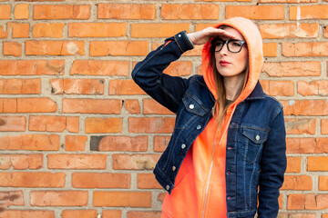 Young girl with glasses, put his hand looking into the distance, wearing a hoodie and denim jacket on a red brick wall