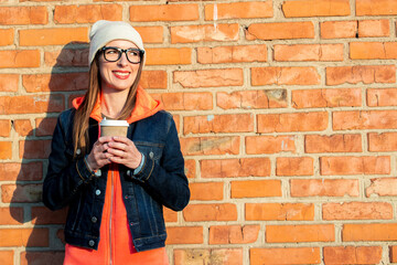 Smiling young girl in glasses holds a paper cup with coffee, looks to the side, is dressed in a bright hat, in a sweatshirt and denim jacket on a background of a red brick wall