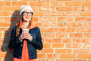 Young woman looks away holds a paper cup with coffee on a background of a brick wall wearing a white hat and a denim jacket.