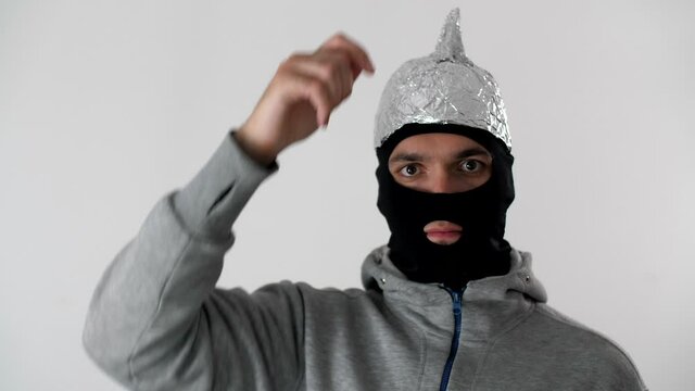 Portrait of a white man  in a black balaclava puts on a foil hat. 5G tower radiation protection. Irrational fear of a non-existent problem. Protective tinfoil helmet to the brains. Guy with a tinfoil 