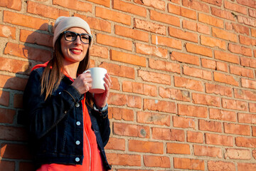 Young smiling woman in glasses holding a white cup on the background of brick wall wearing a gray cap, hoodie and denim jacket