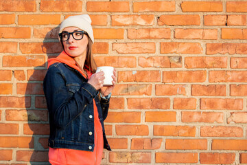 Young woman in glasses holding a white cup on a background of a brick wall, looking back, wearing a gray cap, hoodie and denim jacket. Shot under natural light