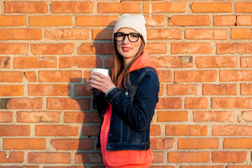 Fototapeta na wymiar Young smiling woman in glasses holding a white cup on a background of a brick wall, wearing a gray cap, hoodie and denim jacket
