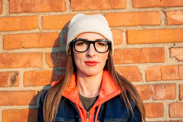 Young woman smiling with glasses against a brick wall, wearing a gray hat, a sweatshirt and a denim jacket, close-up