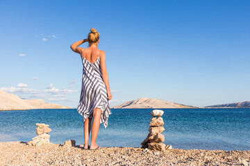 Happy carefree woman wearing beautiful striped summer dress enjoying late afternoon walk on white pabbled beach on Pag island, Croatia.