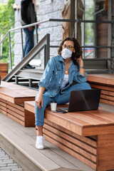 The girl with dark hair in a white medical mask and blue jeans. Young european woman with laptop working outdoors. New life concept for students and freelancers. Pensive serious face