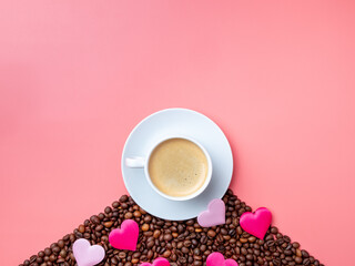 Flat lay. White ceramic cup with coffee stands on a hill of coffee beans on a pink background. Small silk hearts are scattered nearby. Fragrant roasted coffee for breakfast for lovers. Copy space.