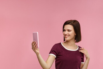 Funny young brunette woman making selfie. Smiling girl wearing t-shirt holding pink smartphone, making faces on camera, posing for selfie isolated on pink background.