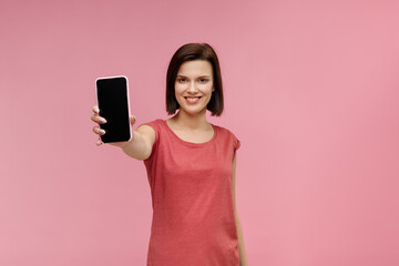 Fototapeta premium Funny brunette woman showing blank screen mobile phone on camera. Young smiling girl demonstrating smartphone display isolated over pink background. Copy space. App for your business.